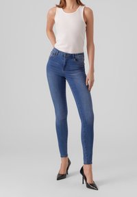 Tmavě modré skinny džíny s vysokým pasem a hladkým denimovým povrchem, spárované s přiléhavým bílým ribbed topem a černými vysokými podpatky.