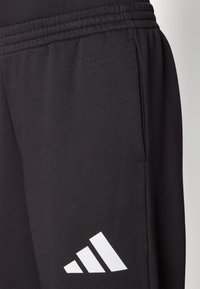 Sort træningsbukser lavet af blødt stof, med elastisk talje, sidelomme og et hvidt Adidas-logo med tre diagonale striber.