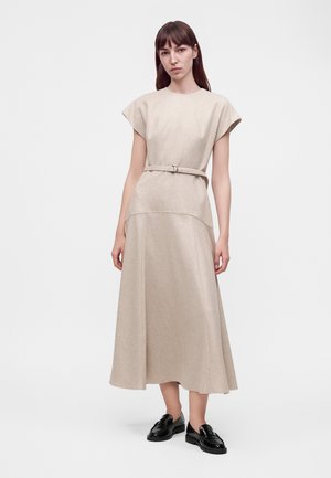Femme aux cheveux bruns raides portant une robe midi beige ceinturée et des mocassins noirs, debout devant un fond clair uni.