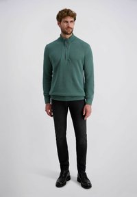 Pullover aus grün gestricktem Stoff mit hohem Kragen und Viertelreißverschluss. Getragen mit eng anliegenden schwarzen Jeans und schwarzen Schnürschuhen.
