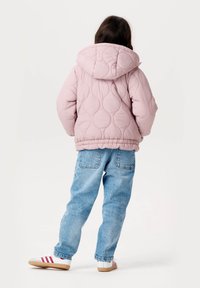 Quiltad rosa pufferjacka med huva, ribbad fåll och sömdetaljer, tillsammans med blå denimjeans och vita sneakers med röda ränder.