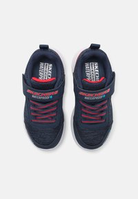Baskets imperméables marine avec des accents rouges, dessus en tissu texturé, lacets élastiques et sangle à fermeture velcro. Semelle blanche et détails de marque.