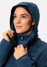 Giacca impermeabile blu scuro con cappuccio, chiusura con cerniera e fodera in pile. Dettagli notevoli includono il girovita regolabile e il logo.