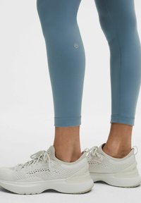 Leggings azules de tela elástica con un diseño ceñido y dobladillo ajustado, combinados con zapatos deportivos color beige claro que presentan una parte superior de malla y suelas gruesas.