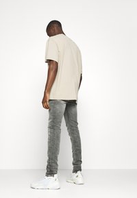 Camiseta beige de manga corta, de corte relajado, combinada con jeans ajustados grises y zapatillas deportivas blancas, de pie sobre un fondo blanco liso.