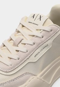 Gros plan sur une sneaker beige Armani Exchange montrant les lacets, les textures en daim et en mesh, ainsi que le logo de la marque sur la languette et le côté.