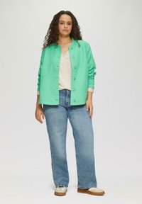 Gepatchte mintgrüne Jacke mit Druckknöpfen, getragen über einem cremefarbenen Oberteil und blauen Jeans. Die Jacke hat lange Ärmel und einen lässigen Schnitt.
