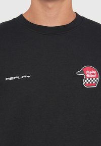 Fekete pamut pulóver bordás nyakkal. A "REPLAY" fehér betűkkel és egy piros-fehér sisak grafikával rendelkezik, amely sakktáblás mintázattal van ellátva.