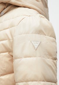 Μπουφάν με επένδυση σε ανοιχτό μπεζ χρώμα, με καπιτονέ σχέδιο. Διαθέτει λευκή τριγωνική τσέπη με λογότυπο στον αριστερό ώμο. Λείο ύφασμα.