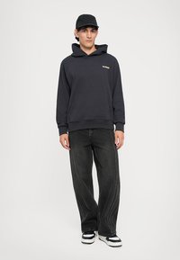 Sudadera negra con bolsillo frontal y logo amarillo, combinada con jeans oscuros de corte amplio que presentan costuras laterales texturizadas y zapatillas.