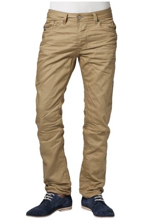 Homme portant un pantalon beige ajusté et des chaussures à lacets en daim bleu foncé, debout les pieds légèrement écartés sur un fond blanc.