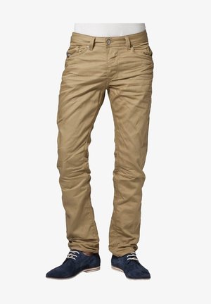Homme portant un pantalon beige ajusté et des chaussures à lacets en daim bleu foncé, debout les pieds légèrement écartés sur un fond blanc.