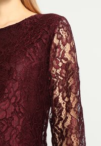 Burgundy spetsblus med långa ärmar, prydd med blommönster och en strukturerad yta. Tyget är känsligt och något genomskinligt.