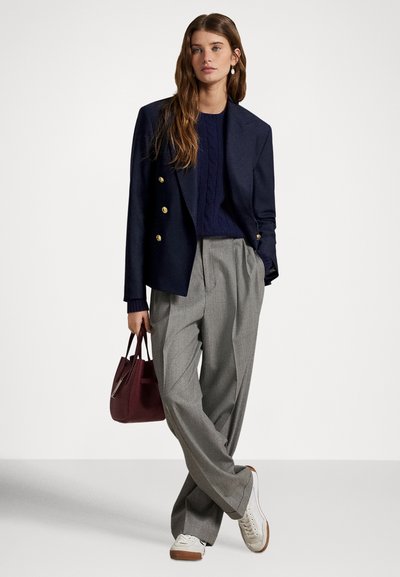 Kobieta w granatowej marynarce na granatowym swetrze, szare szerokie spodnie i białe trampki, trzyma bordową torebkę, jedną ręką w kieszeni.