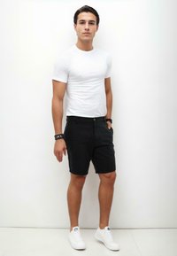 White fitted t-shirt, black shorts with pockets, flat front, and button closure; model wears white sneakers; plain background.