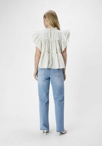 Blusa bianca di cotone con maniche a volant, colletto alto e vita elastica. Indossata sopra jeans a gamba dritta blu con una texture sbiadita.