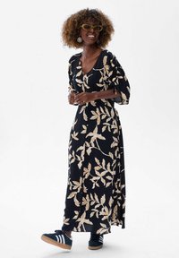 Zwarte maxi-jurk met een beige bladpatroon, v-hals en pofmouwen. Gecombineerd met zwart-witte sneakers. Glad materiaal, relaxte pasvorm.