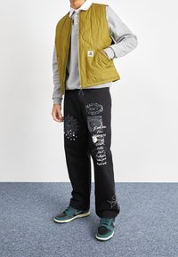 Olivegrön kviltad väst över en grå sweatshirt, tillsammans med svarta byxor med vita grafiska tryck. Svart och teal sneakers fulländar looken.