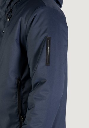 Giacca trapuntata blu navy con finitura testurizzata, dotata di tasca laterale con zip e patch con logo sulla manica. Design con cappuccio.