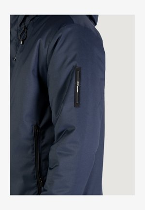 Giacca trapuntata blu navy con finitura testurizzata, dotata di tasca laterale con zip e patch con logo sulla manica. Design con cappuccio.
