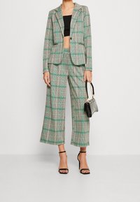 Femme portant un pantalon large à carreaux verts et noirs et un blazer assorti, un crop top noir, des talons noirs, et tenant un sac à main noir.