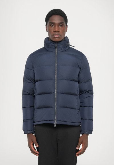 Veste matelassée bleu marine avec col montant, fermeture éclair à l'avant et design matelassé. Tissu lisse avec des coutures horizontales et un matériel minimal.