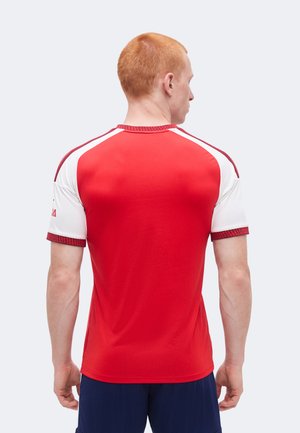 Homme aux cheveux courts rouges portant un maillot de sport rouge et blanc et un short bleu marine, tourné de dos à la caméra sur un fond uni.
