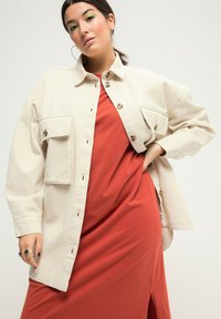 Beige button-up jas met twee borstzakken over een oranje jurk. De jas heeft een relaxte pasvorm, zichtbare stiksels en grote knopen.