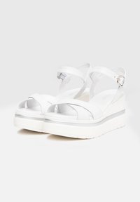 Sandales à plateforme blanches avec des brides croisées, boucles réglables et semelle texturée. Présente un léger accent argenté sur la semelle intermédiaire.