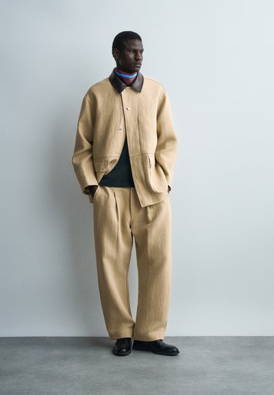 Άνδρας φορώντας μπεζ υπερμεγέθη σακάκι και αντίστοιχο φαρδύ παντελόνι, μαύρα παπούτσια και πολύχρωμο ζιβάγκο, στέκεται μπροστά σε απλή φωτεινή τοίχο.