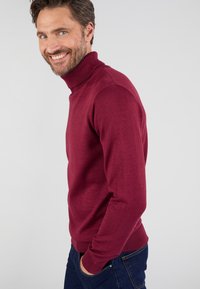 Glunderende man met een baard en blauwe ogen, gekleed in een bordeauxrode coltrui en donkere spijkerbroek, staat voor een effen achtergrond.