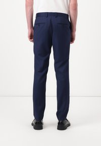 Pantalones azul marino con un corte ajustado, que cuentan con dos bolsillos traseros y trabillas para cinturón. Fabricados con una tela suave y ligera. Zapatos negros visibles.