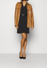 Žena oblečená v černých šatech s páskem, s hnědou koženou bundou na knoflíky a černými lodičkami s špičatou špičkou, stojící na pozadí s bílou plochou.