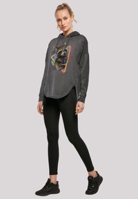 Grå hettegenser med et grafisk trykk av en vaskebjørn og fargerike geometriske detaljer, kombinert med svarte leggings og svarte joggesko.