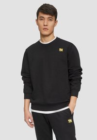 Sweatshirt noir en coton avec un col rond et des poignets côtelés. Il présente un petit patch logo jaune sur la poitrine gauche.