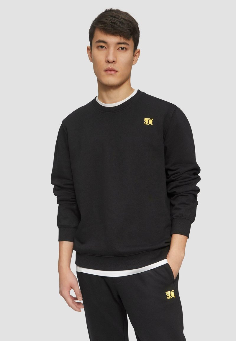 Sweatshirt noir en coton avec un col rond et des poignets côtelés. Il présente un petit patch logo jaune sur la poitrine gauche.