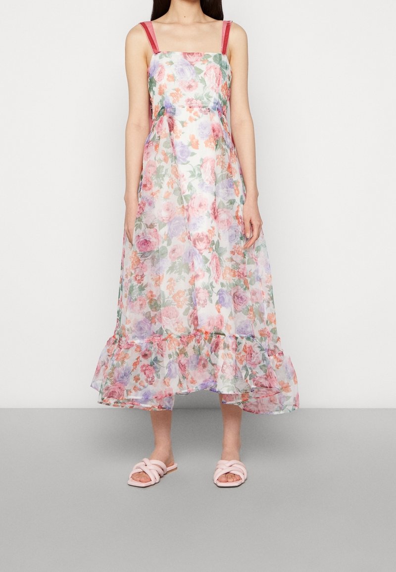 Femme portant une robe midi sans manches fleurie avec un ourlet volanté rose et des bretelles rouges, associée à des sandales légères rose clair.
