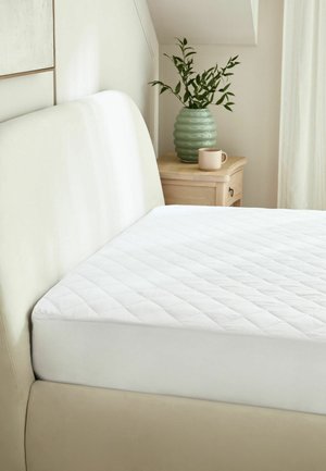 Beiges gepolstertes Bett mit weißem, gestepptem Matratzenschoner, neben einem hölzernen Nachttisch mit grüner Vase mit Blättern und einer beigen Kaffeetasse.
