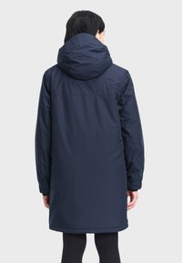 Marineblauwe, knie-lange parka met een grote capuchon, elastische manchetten en een lichtgewicht, waterafstotend materiaal. Strakke lijnen en minimale details.