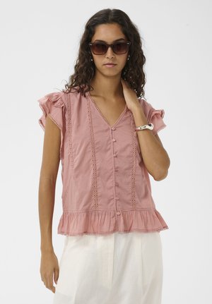 Mujer con cabello ondulado que lleva gafas de sol marrones, una blusa rosa sin mangas con volantes y botones, y pantalones blancos, tocándose el cuello con una mano.