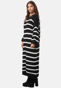 Gestreiftes Strickkleid in Schwarz-Weiß mit lockerer Passform, langen Ärmeln und seitlichen Taschen, ergänzt durch schwarze High-Heels-Stiefel.