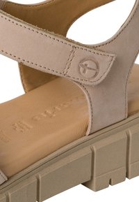 Beige suede sandal featuring a textured strap, reinforced stitching, and a light brown insole. The sole is chunky and textured for grip.
