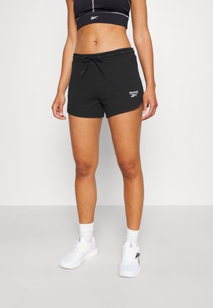 Woman wearing black Reebok drawstring shorts, black sports bra, white socks, and white sneakers standing against a plain background.