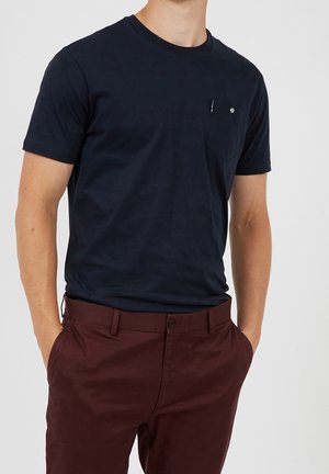 Uomo che indossa una semplice t-shirt a maniche corte blu scuro e pantaloni rosso scuro con le mani in tasca su uno sfondo bianco.
