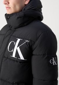 Veste matelassée noire avec un col montant, un logo "CK" blanc bien visible à l'avant et un écusson de marque sur la manche.