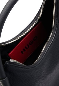 Schwarze Lederhandtasche mit Reißverschluss, die ein rotes Innenfach mit "HUGO"-Branding und einer strukturierten Oberfläche zeigt.
