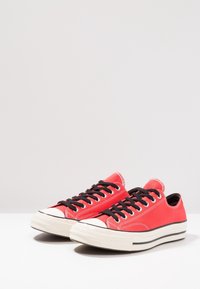 Baskets en toile rouges à tige basse avec des lacets noirs, embouts en caoutchouc blanc et semelles blanches avec des rayures noires sur une surface blanche.