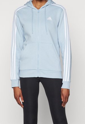 Personne portant un sweat à capuche zippé Adidas bleu clair avec des bandes blanches sur les manches et un legging noir, visible du cou aux hanches.