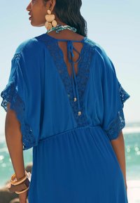 Blauwe kaftan jurk met kantaccenten, korte mouwen, een open rug en een striksluiting. Accessoires zijn grote statement oorbellen en houten armbanden.