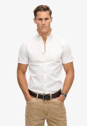 Korte mouwen, wit overhemd met kraag en knoopsluiting, voorzien van een borstzak; gedragen met beige broek en een zwarte riem, aangevuld met een horloge.